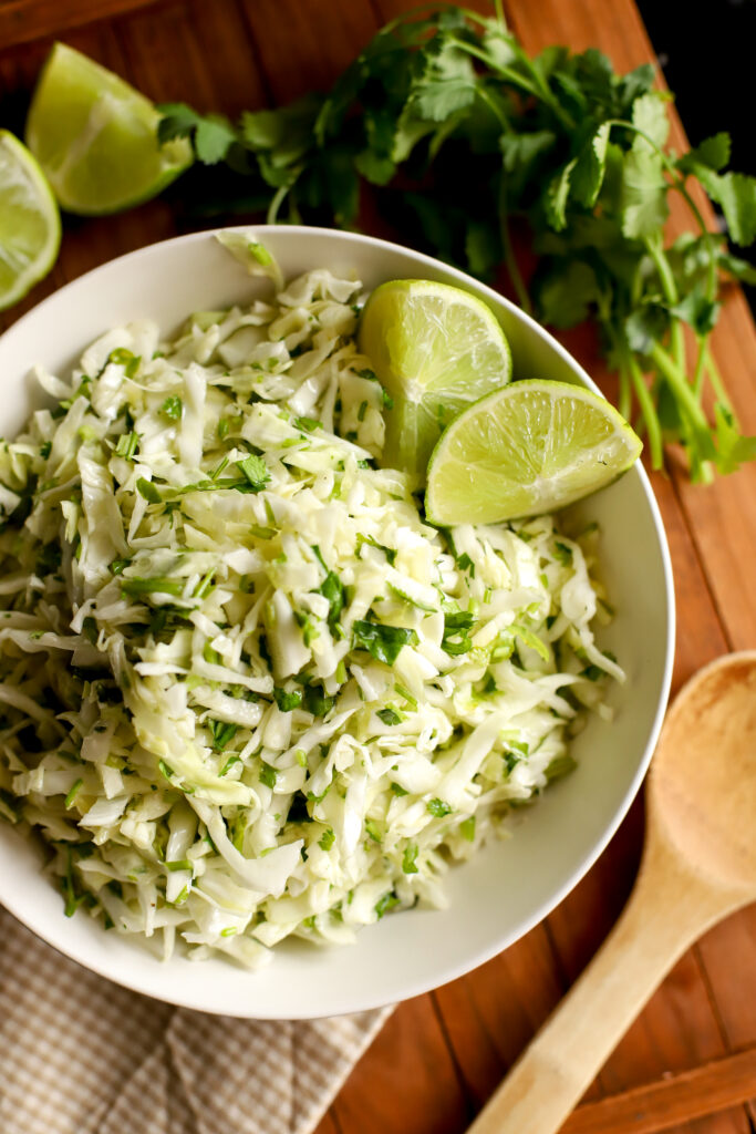 up close shot of cilantro lime cabbage slaw in a bowl with limes and cilantro garnish