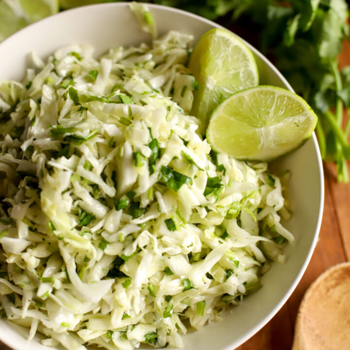 an up close shot of cilantro lime cabbage slaw in a bowl with sliced limes as garnish