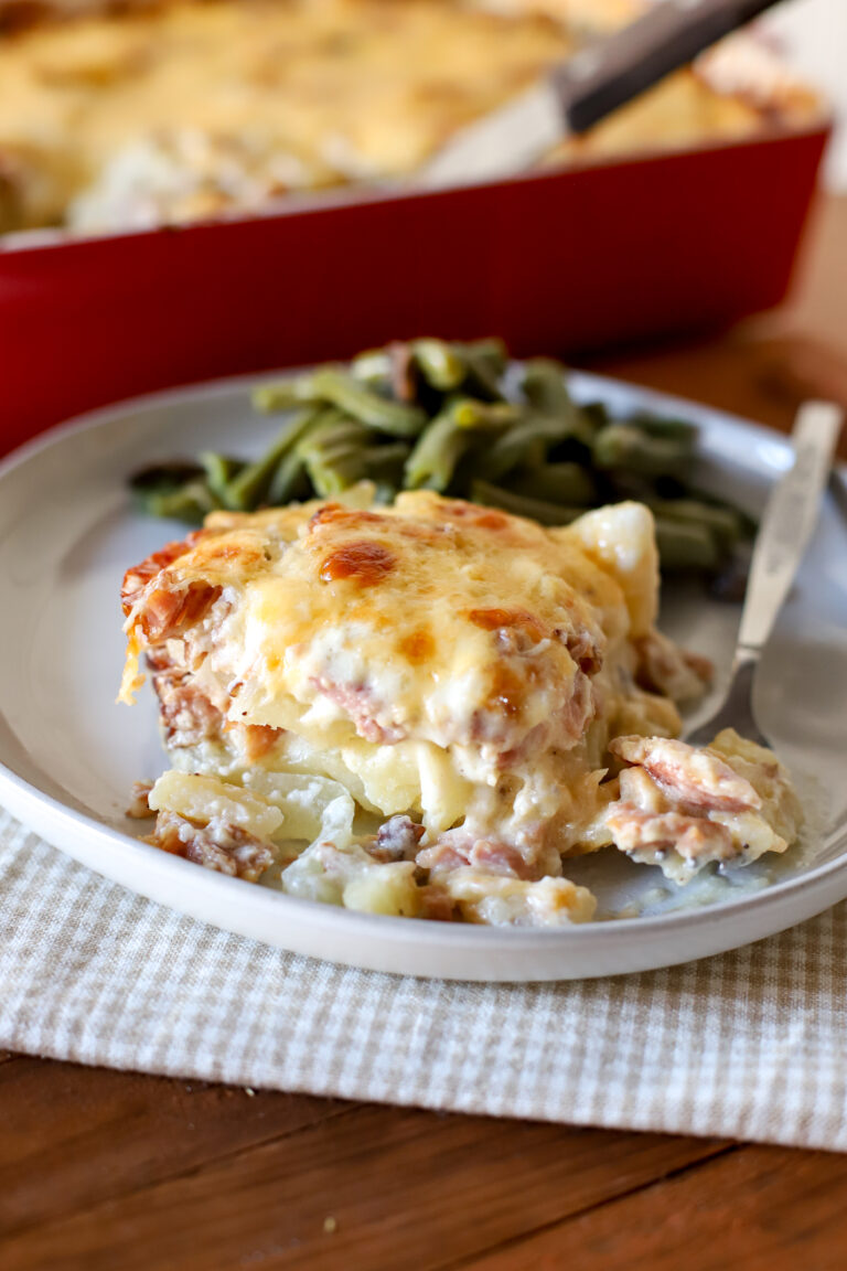 up close shot of a slice of scalloped potatoes and ham being served with green beans and a pan of scalloped potatoes and ham in the background