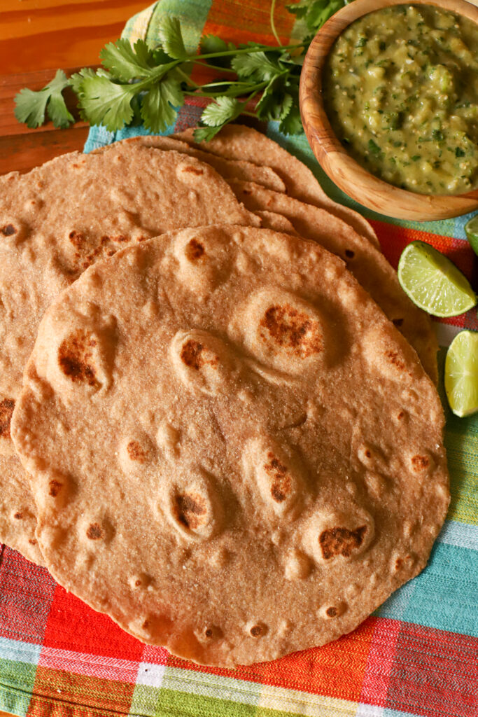 up close shot of stack of fresh milled tortillas on a checkered towel with salsa, cilantro, and sliced limes