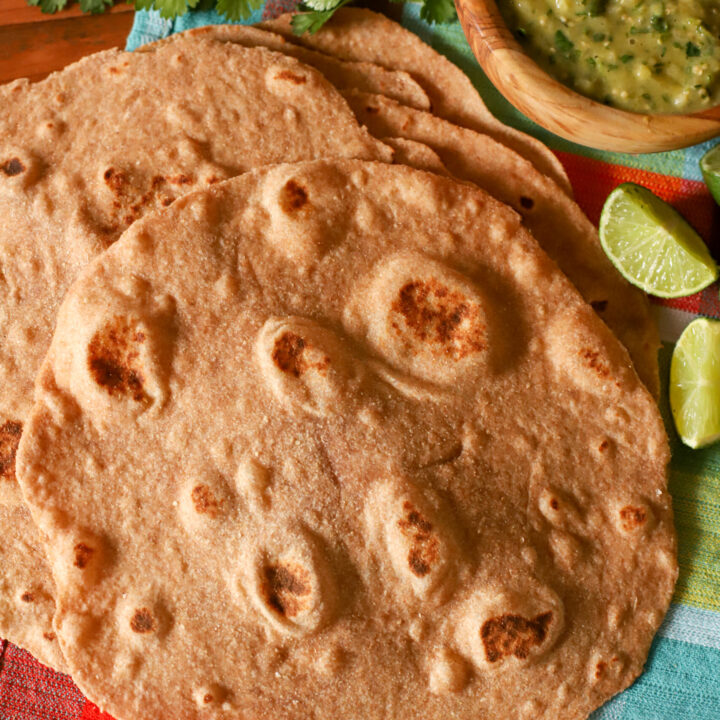 up close shot of stack of fresh milled tortillas on a checkered towel with salsa, cilantro, and sliced limes