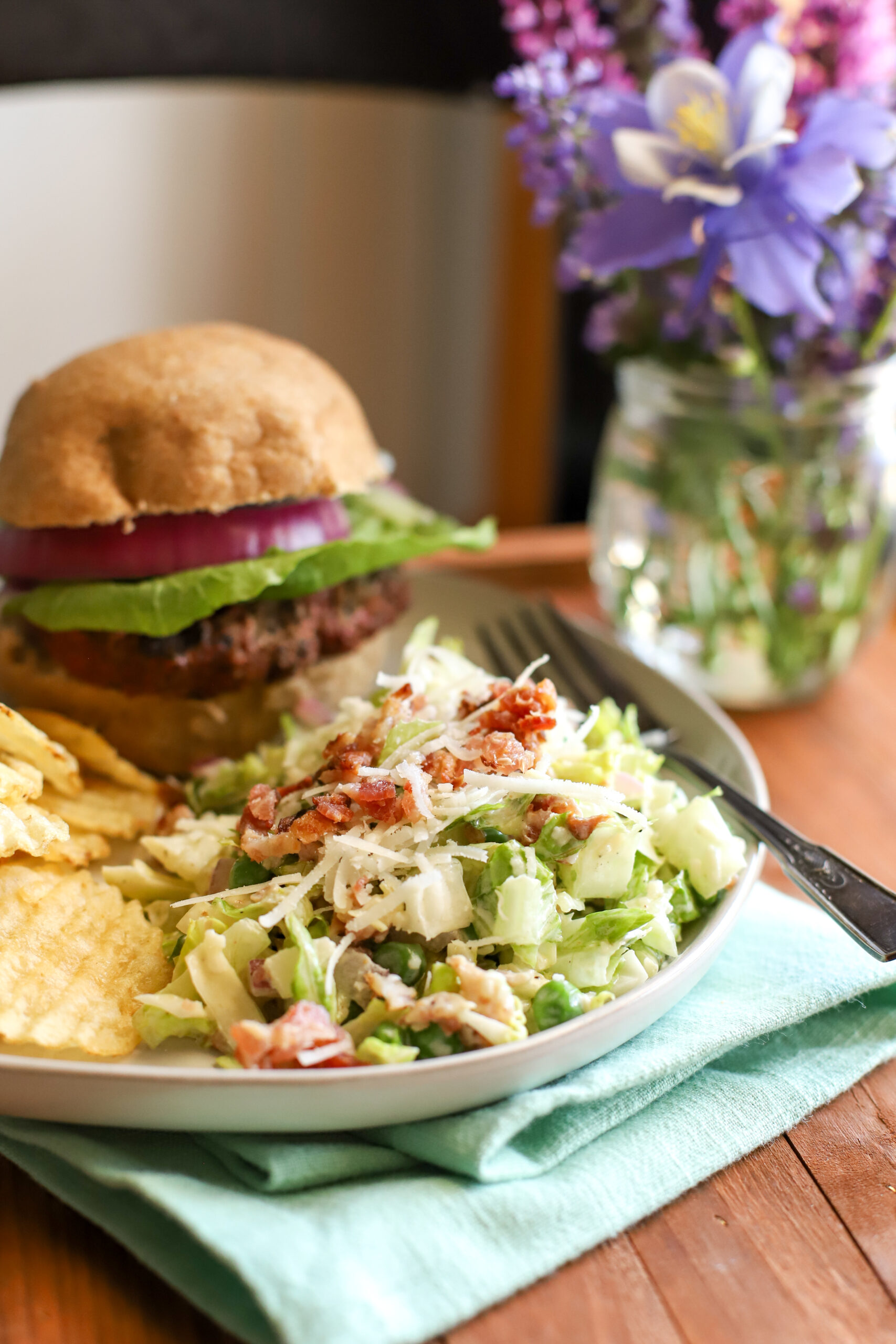 an up close shot of 7 layer salad on a plate with a hamburger and chips placed on a teal blue towel with a vase of flowers in the background